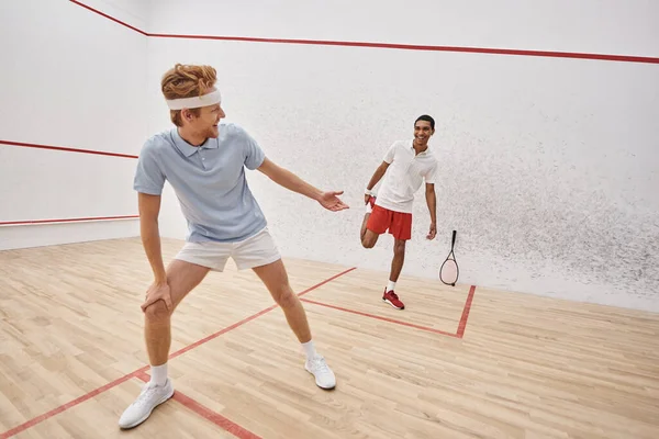 Happy redhead man with headband doing lunges near african american friend inside squash court — Stock Photo