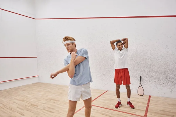 Young redhead man with headband warming up near happy african american friend inside squash court — Stock Photo