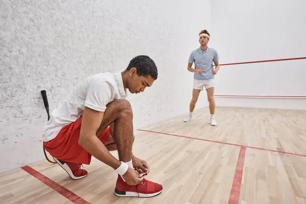 African american man tying shoelaces while sitting inside of squash court near redhead friend — Stock Photo