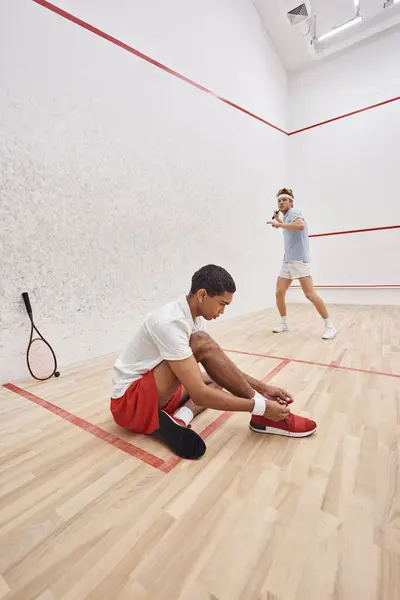 African american man tying shoelaces while sitting near redhead friend inside of squash court — Stock Photo