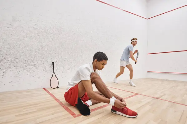African american player tying shoelaces while sitting near redhead friend inside of squash court — Stock Photo