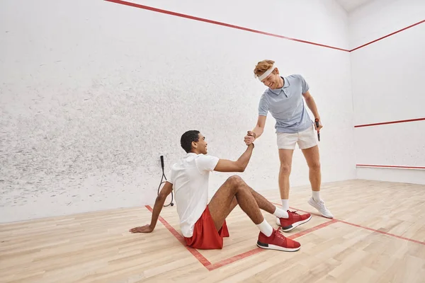 Feliz ruiva homem ajudando seu amigo afro-americano a levantar-se do chão dentro da corte de squash — Fotografia de Stock