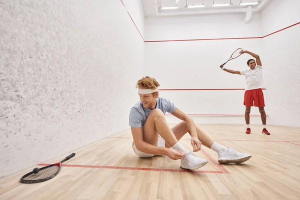 Happy man tying shoelaces while sitting near african american friend inside of squash court — Stock Photo
