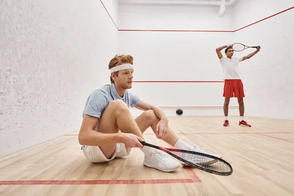 Hombre en ropa deportiva celebración de raqueta mientras está sentado cerca de amigo afroamericano dentro de la cancha de squash - foto de stock