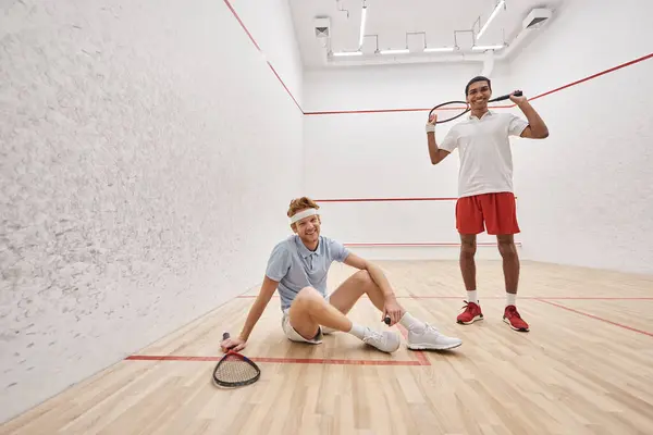 Happy man holding racquet while sitting near african american friend inside of squash court — Stock Photo
