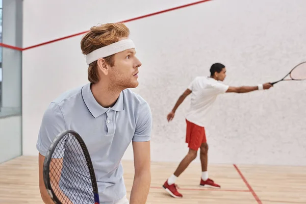 Homem ruivo em sportswear e headband jogando com amigo afro-americano dentro da corte de squash — Fotografia de Stock