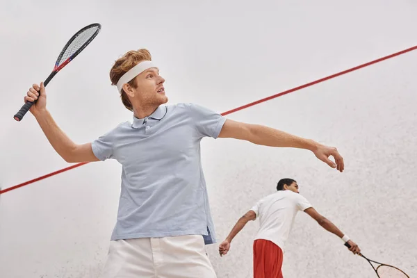 Rothaariger Mann in Sportbekleidung und Stirnband spielt mit afrikanisch-amerikanischem Freund im Squash-Court — Stockfoto