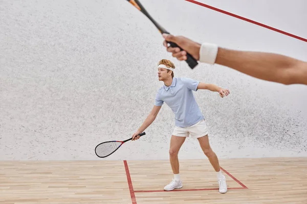Redhead man in sportswear and headband playing with african american friend inside of squash court — Stock Photo