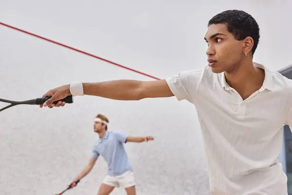 Enfoque en el hombre afroamericano en ropa deportiva jugando con un amigo borroso dentro de la cancha de squash - foto de stock