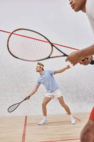 Concentre-se no homem ruivo em sportswear brincando com amigo afro-americano dentro da corte de squash — Fotografia de Stock