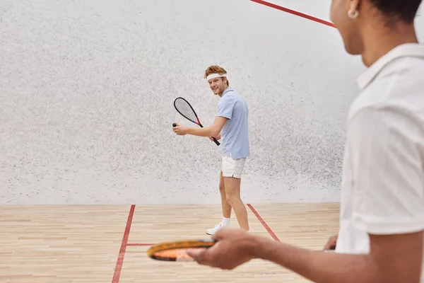 Se concentrer sur l'homme heureux en vêtements de sport jouer avec un ami afro-américain à l'intérieur de court de squash — Photo de stock