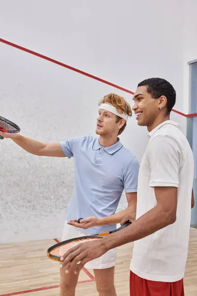 Jogadores inter-raciais felizes em sportswear sorrindo e olhando para o interior da quadra de squash, raquetes — Fotografia de Stock
