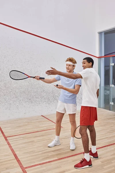 Happy interracial male friends pointing with racquets and looking away inside of squash court — Stock Photo