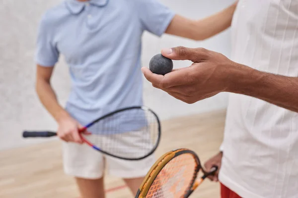 Vista recortada del hombre afroamericano sosteniendo pelota de squash cerca de amigo dentro de la cancha, jugadores - foto de stock