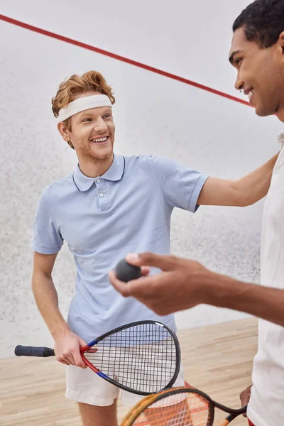 Happy african american man holding squash ball near redhead friend inside of court, players — Stock Photo