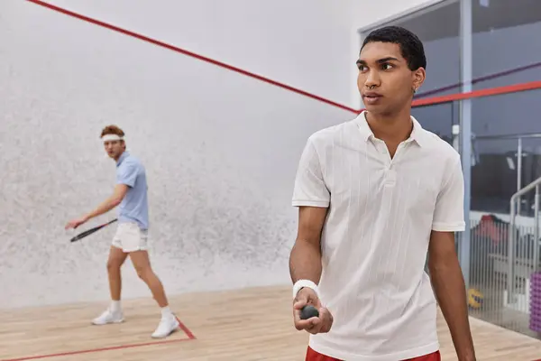 Young african american man holding squash ball and playing with redhead friend inside of court — Stock Photo