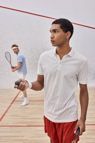 Guapo afroamericano hombre sosteniendo pelota de squash y jugando con pelirroja amigo dentro de la corte - foto de stock