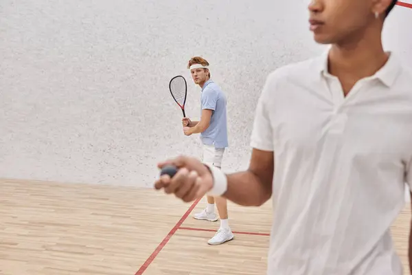 Pelirroja sosteniendo raqueta cerca de hombre afroamericano con pelota de squash jugando en corte — Stock Photo