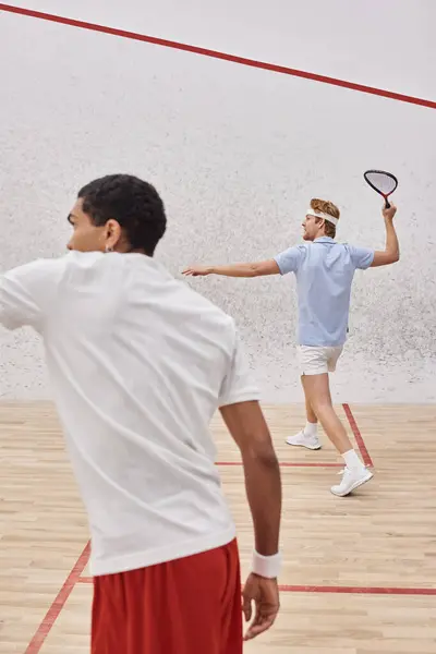 Ruiva homem segurando squash raquete perto de homem americano africano enquanto jogando juntos na corte — Fotografia de Stock