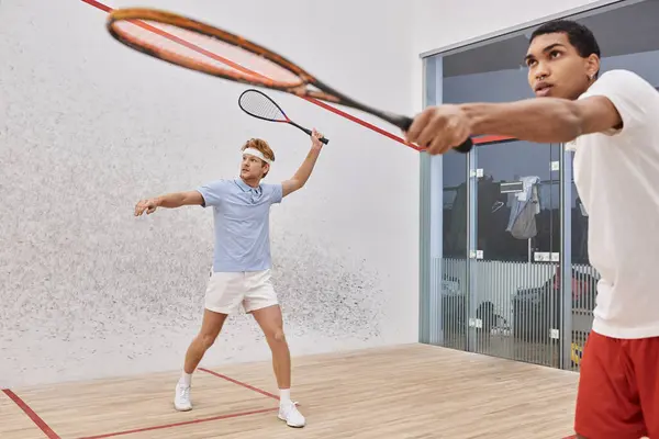 Jogador ruiva segurando raquete squash perto de homem americano africano enquanto jogam juntos na corte — Fotografia de Stock