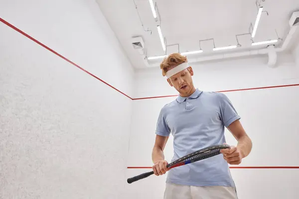 Homme fatigué en bandeau et vêtements de sport respirant lourdement et regardant la raquette de squash à l'intérieur de la cour — Photo de stock
