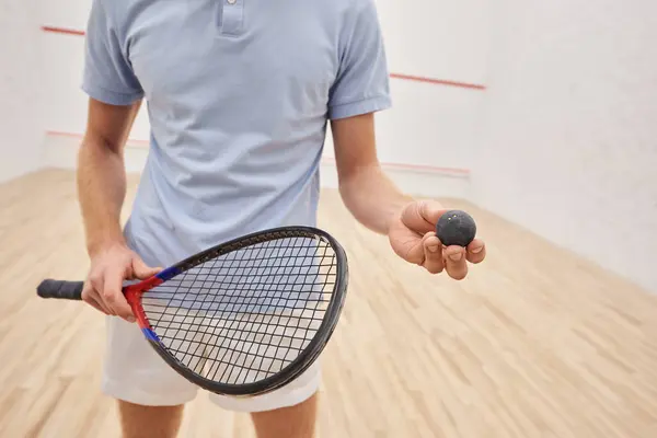 Обрезанный вид человека в спортивной одежде держа мячик для сквоша и ракетку и стоя внутри корта — стоковое фото