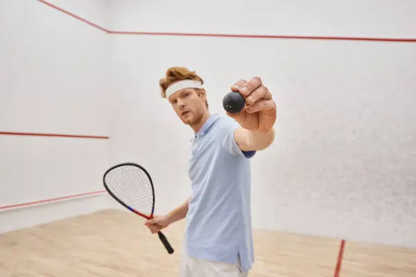 Redhead man in sportswear showing squash ball and holding racquet while standing inside of court — Stock Photo