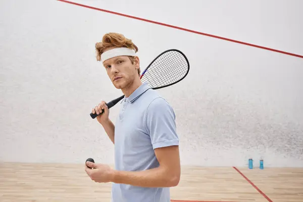 Pelirrojo en ropa deportiva sosteniendo pelota de squash y raqueta mientras está de pie dentro de la cancha cubierta — Stock Photo