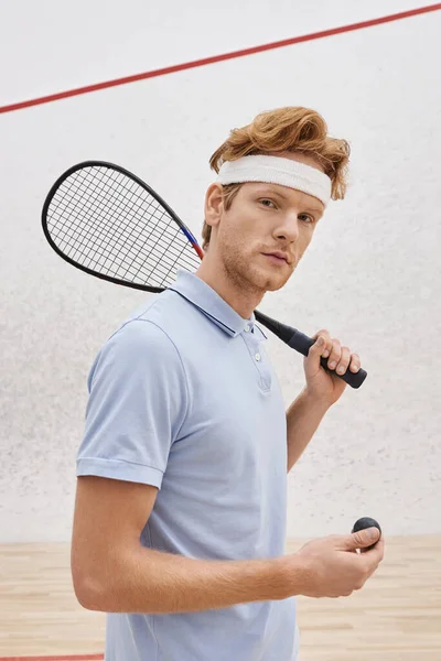 Pelirrojo en ropa deportiva sosteniendo pelota de squash y raqueta mientras está de pie dentro de la cancha, vertical — Stock Photo
