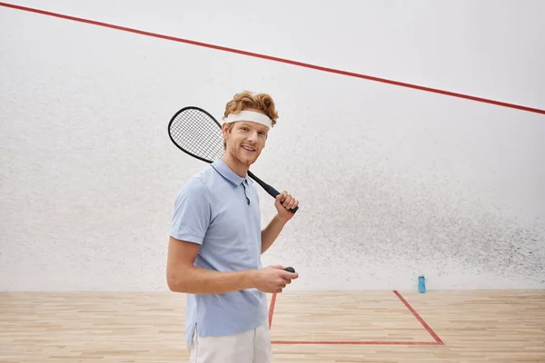 Homem feliz em sportswear segurando bola de squash e raquete enquanto está dentro da quadra interna — Fotografia de Stock