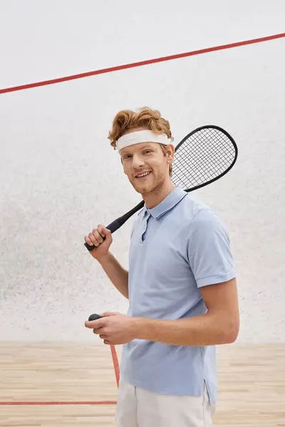 Uomo felice in abbigliamento sportivo in possesso di palla da squash e racchetta mentre in piedi all'interno della corte, verticale — Foto stock