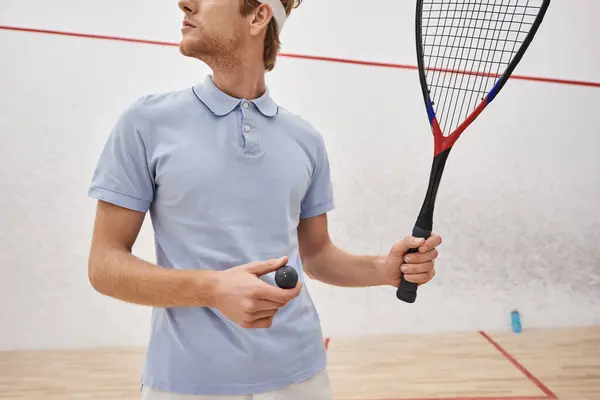 Обрізаний знімок людини в спортивному одязі, що тримає м'яч для сквошу та ракетку, стоячи всередині двору — стокове фото