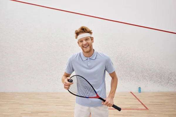 Положительный спортсмен в спортивной одежде держа сквош мяч и ракетку, стоя внутри корта — стоковое фото
