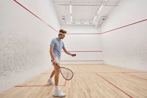 Pelirroja deportista en uso activo sosteniendo pelota de squash y raqueta mientras juega dentro de la cancha - foto de stock
