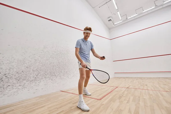 Desportista alegre no desgaste ativo segurando bola de squash e raquete enquanto joga dentro do tribunal — Fotografia de Stock