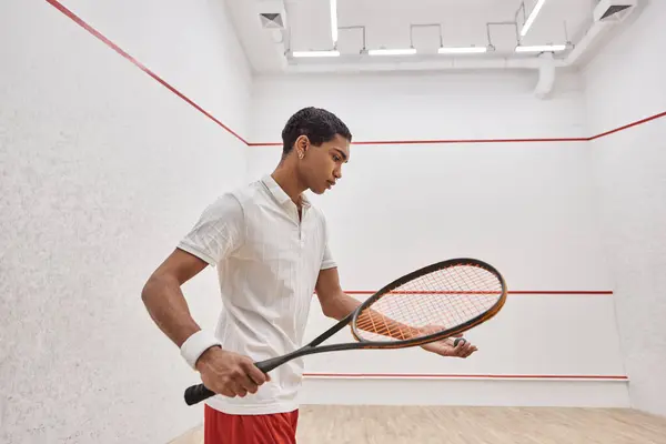 Homem americano africano em desgaste ativo segurando bola de squash e raquete enquanto joga dentro da quadra — Fotografia de Stock
