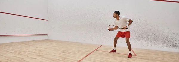 Banner de vista lateral, atlético afroamericano hombre sosteniendo raqueta de squash mientras juega juego en la corte - foto de stock
