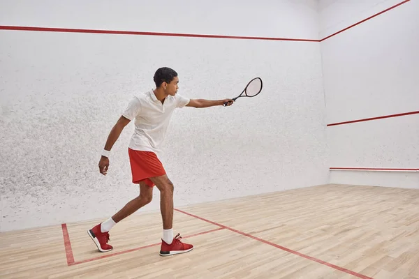 Vista lateral, atlético afro-americano em desgaste ativo segurando raquete enquanto joga squash — Fotografia de Stock