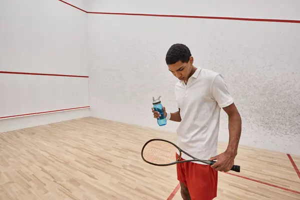 Athletic african american sportsman holding bottle of water and squash racquet after playing game — Stock Photo