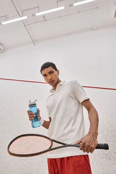 Jeune sportif afro-américain tenant une bouteille d'eau et une raquette de squash après avoir joué au jeu — Photo de stock