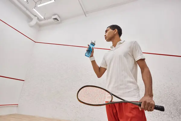Low angle, african american sportsman holding bottle of water and squash racquet after playing game — Stock Photo