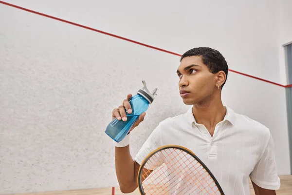 Молодой африканский американец держит в руках спортивную бутылку воды и ракетку для сквоша после игры — стоковое фото