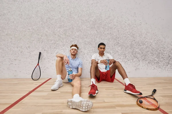 Two tired interracial friends with squash racquets and bottle with water sitting on floor in court — Stock Photo