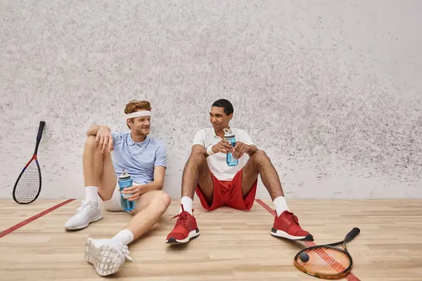 Felizes amigos inter-raciais com squash racquets e garrafa com água sentada no chão no tribunal — Fotografia de Stock