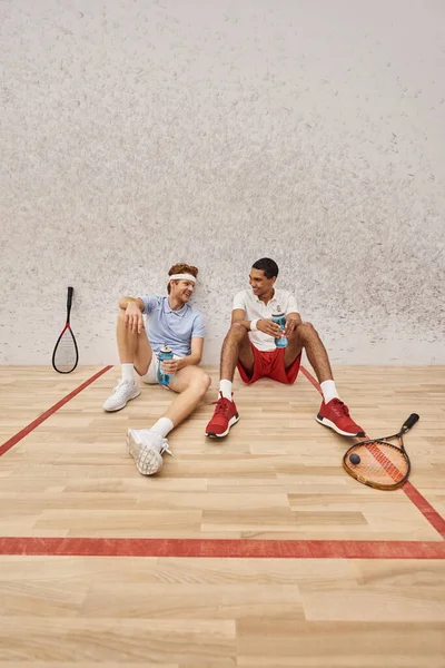 Cheerful interracial friends with squash racquets and bottle with water sitting on floor in court — Stock Photo