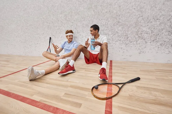 Веселі міжрасові спортсмени з сквошу ракетки і пляшка з водою, сидячи на підлозі в суді — стокове фото