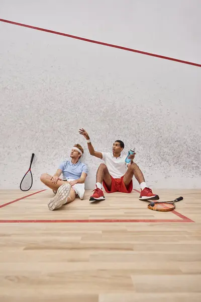 Dois desportistas inter-raciais com raquetes de squash e garrafa com água sentada no chão no tribunal — Fotografia de Stock