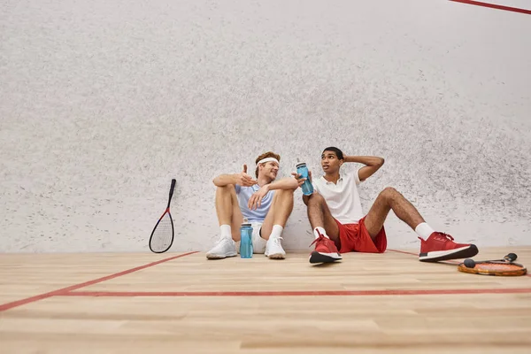 Dos deportistas interracial con raquetas de squash y botellas con agua sentados en el suelo dentro de la cancha - foto de stock