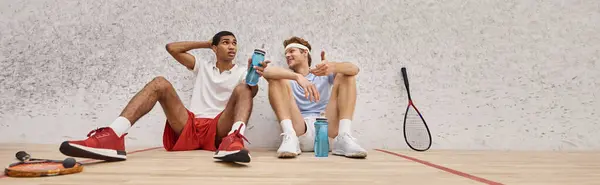 Banner, interracial sportsmen with squash racquets and bottles with water sitting on floor in court — Stock Photo