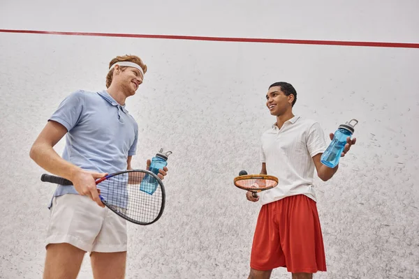 Desportistas inter-raciais felizes com raquetes de squash e garrafas com água em pé dentro do tribunal — Fotografia de Stock